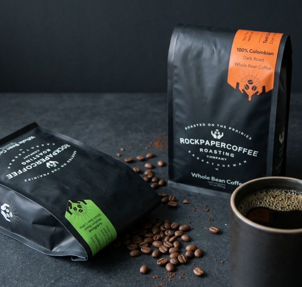 Rock Paper Coffee bags beside a cup of coffee on a dark tabletop